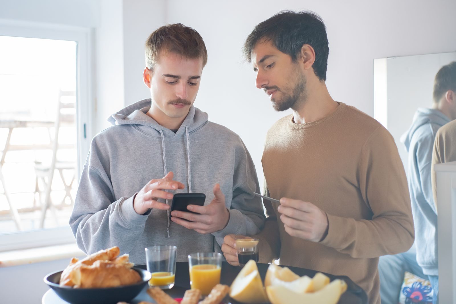 Two young men enjoy a breakfast in Portugal using a smartphone indoors, highlighting a relaxed lifestyle.