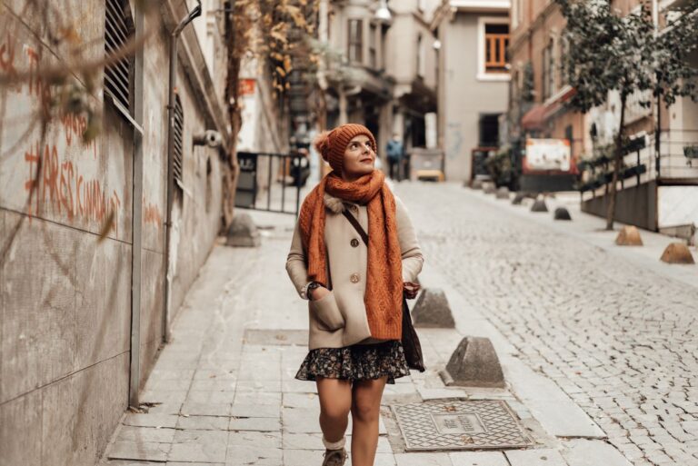 Young female in warm coat scarf and hat with bag looking up while walking on paved street near building in daytime in autumn
