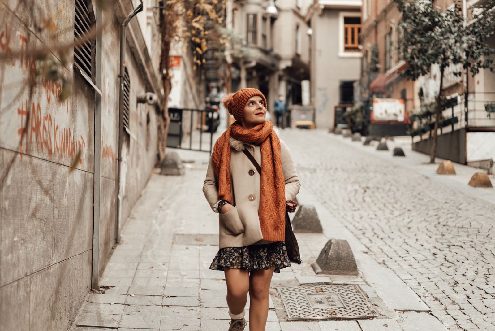 Young female in warm coat scarf and hat with bag looking up while walking on paved street near building in daytime in autumn