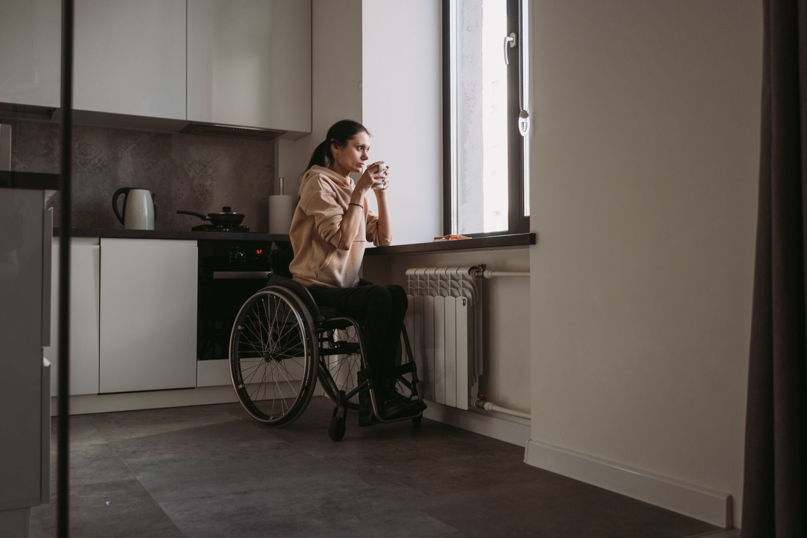 A woman in a wheelchair holding a coffee cup, sitting indoors by a window.