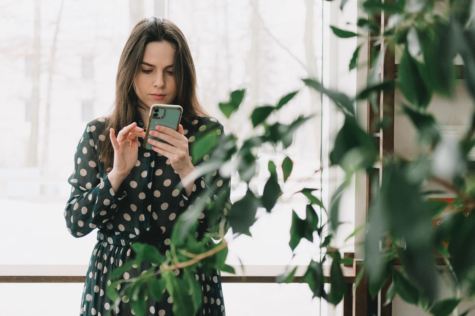 Woman in polka dot dress using smartphone indoors, surrounded by plants.