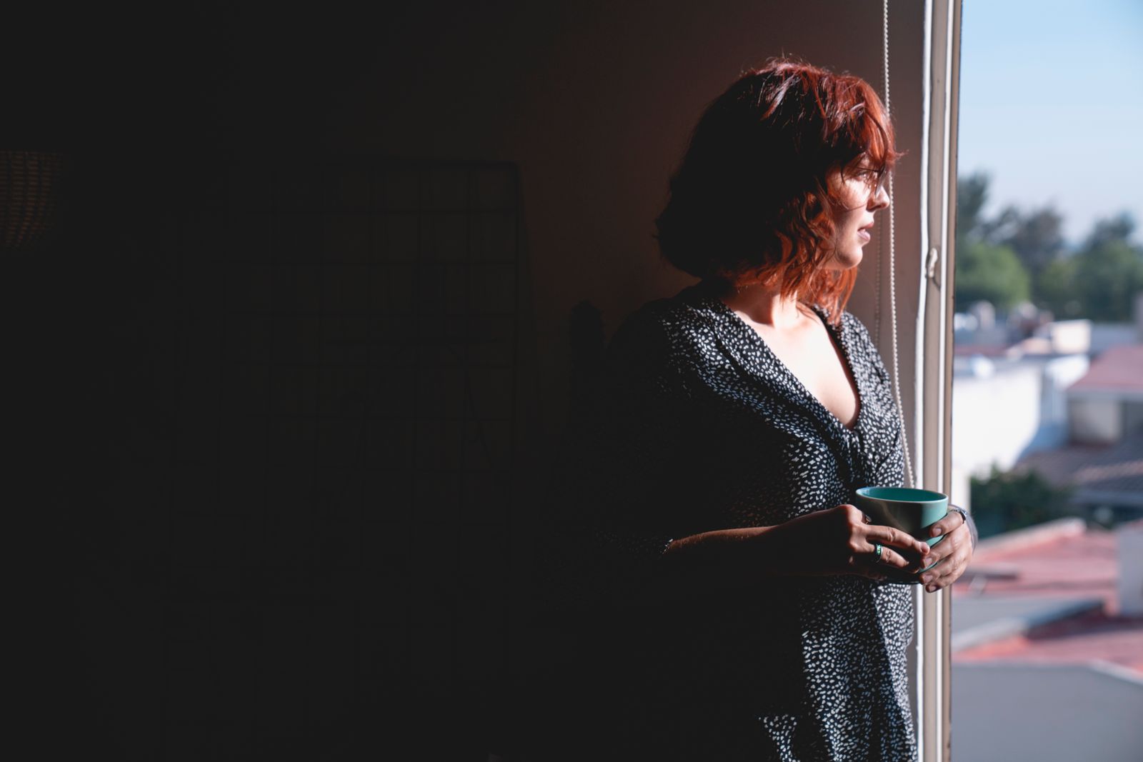 A woman stands by a window, holding a mug and lost in thoughtful contemplation.