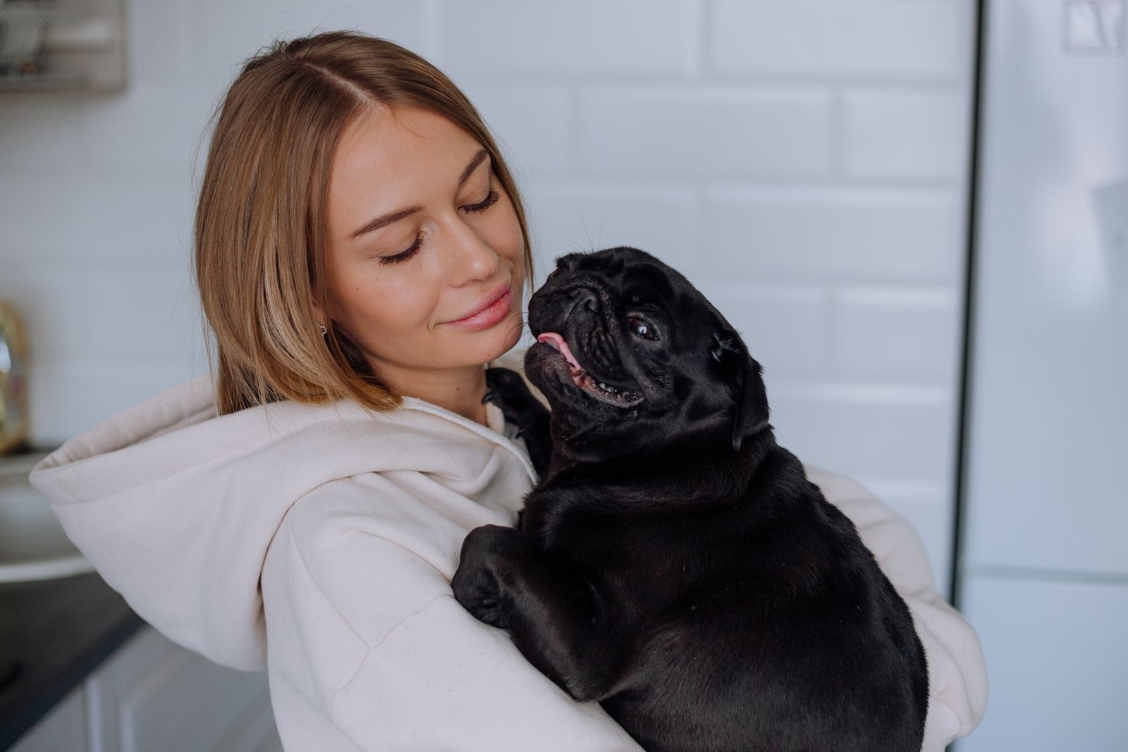 A woman in a hoodie lovingly holds a black pug indoors, showcasing warmth and pet affection.