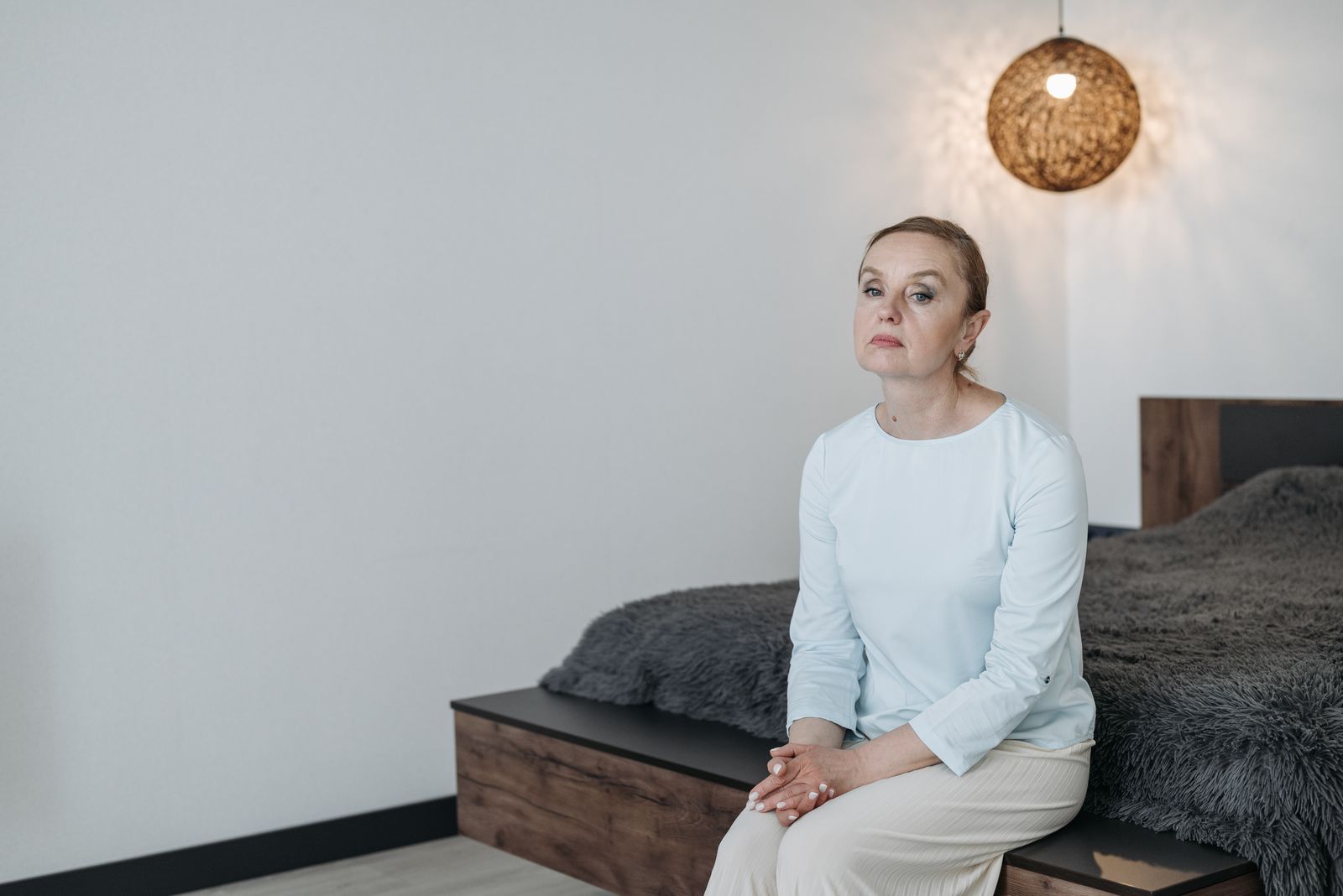 Middle-aged woman sitting on a bed in a modern bedroom, looking thoughtful.