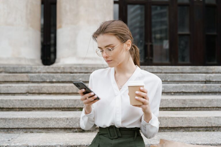 Young woman in business attire using smartphone while enjoying a coffee outdoors.