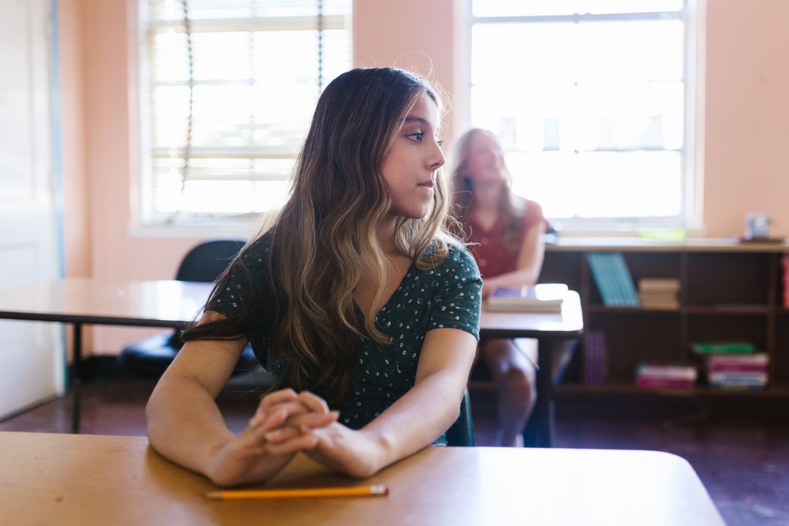 A student sitting thoughtfully in a sunlit classroom with bookshelves in the background.