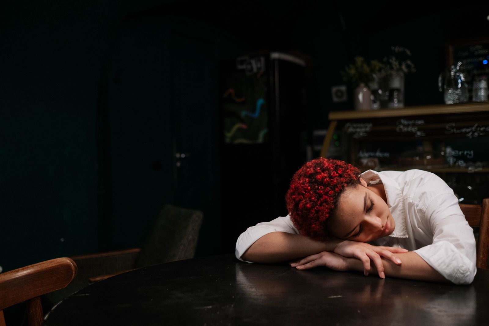 A woman with red hair peacefully resting on a table in a dimly lit cafe.
