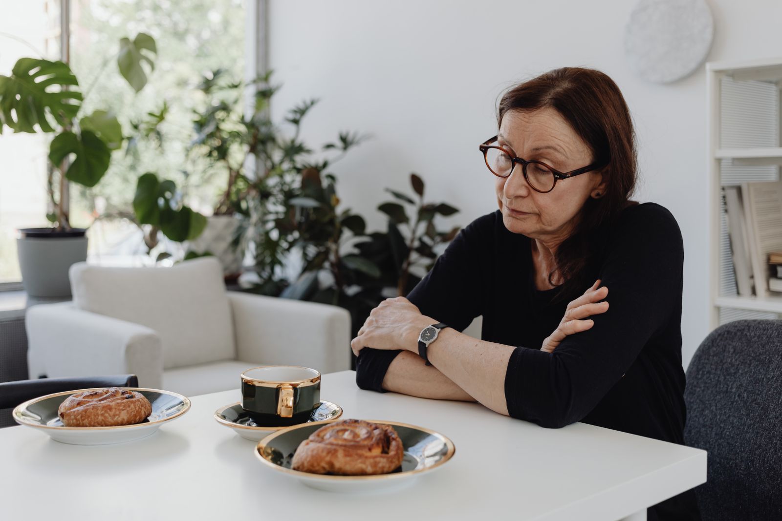 Middle-aged woman in glasses sits at breakfast table with pastries, contemplating.