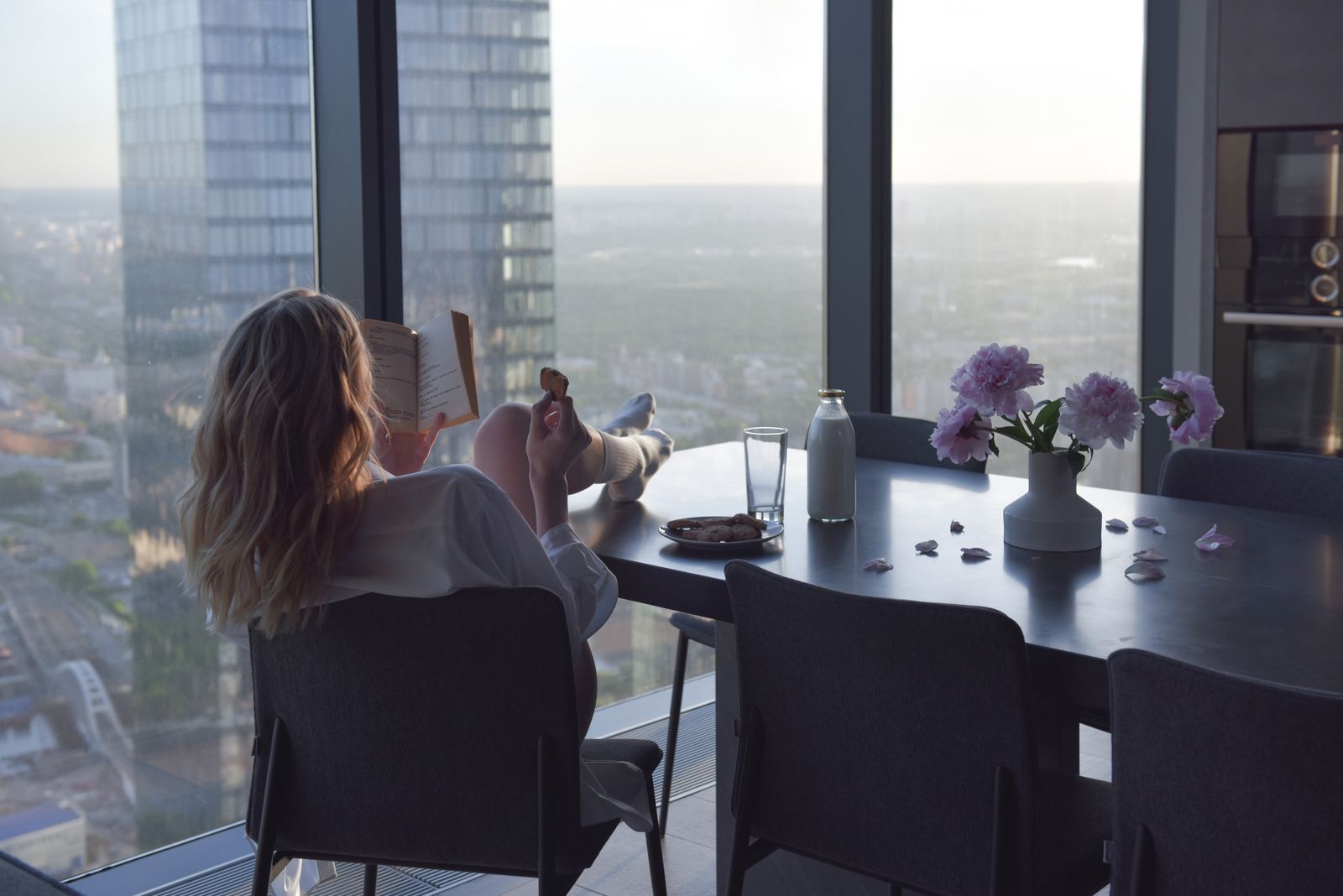 Woman enjoying a book by a window with city view, in a modern indoor setting.