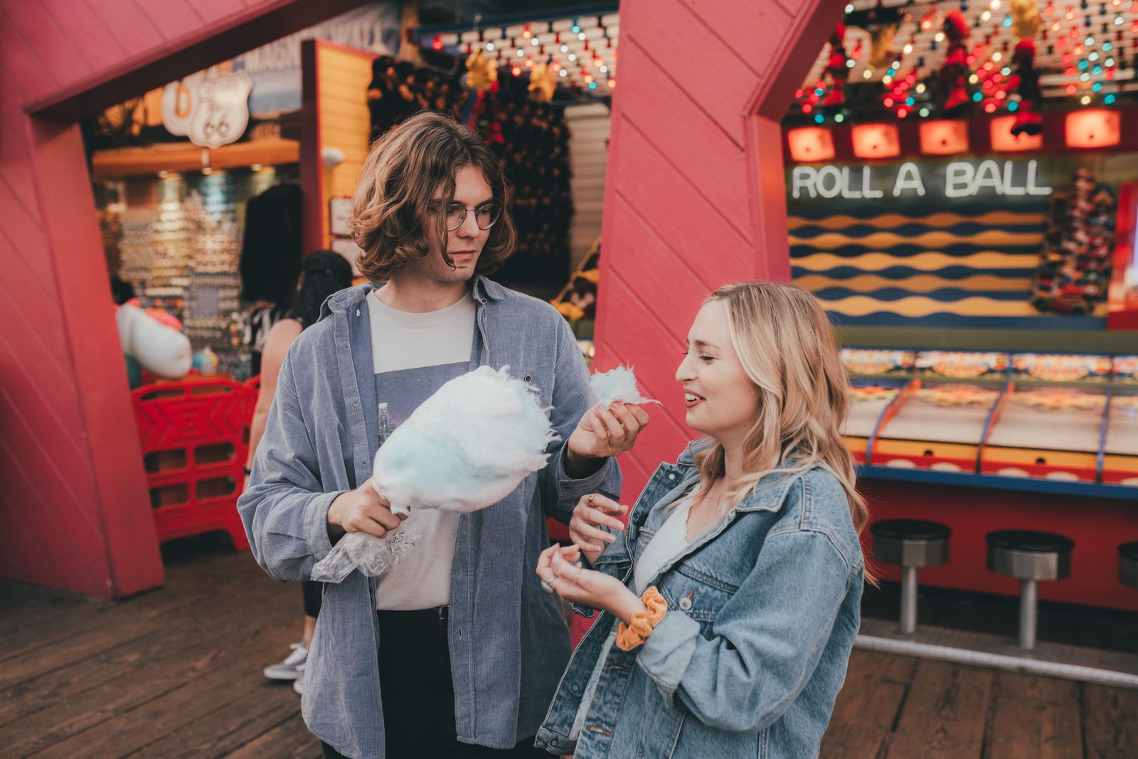 A young couple sharing cotton candy at a lively amusement park game stall.