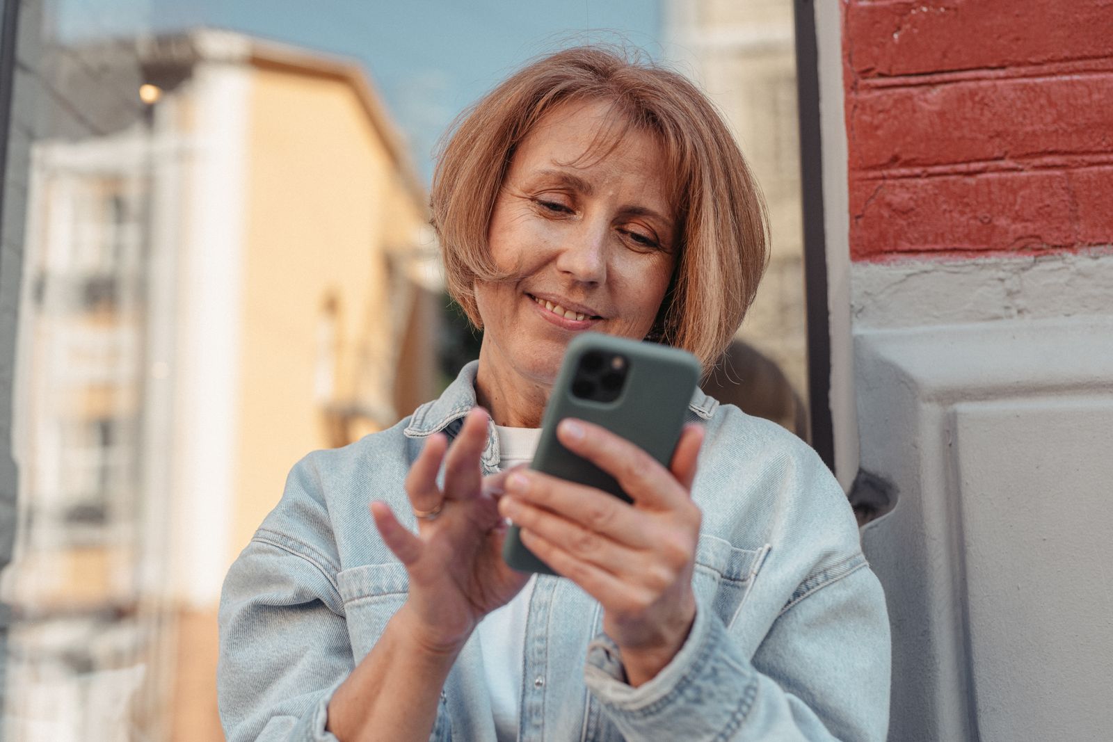 A middle-aged woman in a denim jacket smiles while using her smartphone outside.