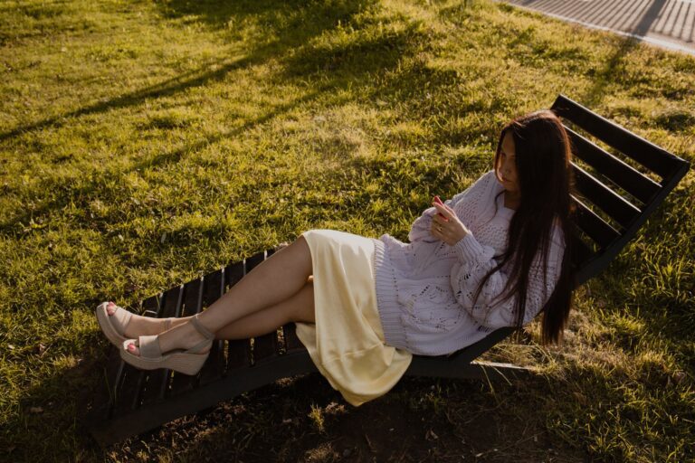 A woman in a sweater relaxes on a lounge chair in a grassy park, enjoying a sunny day.