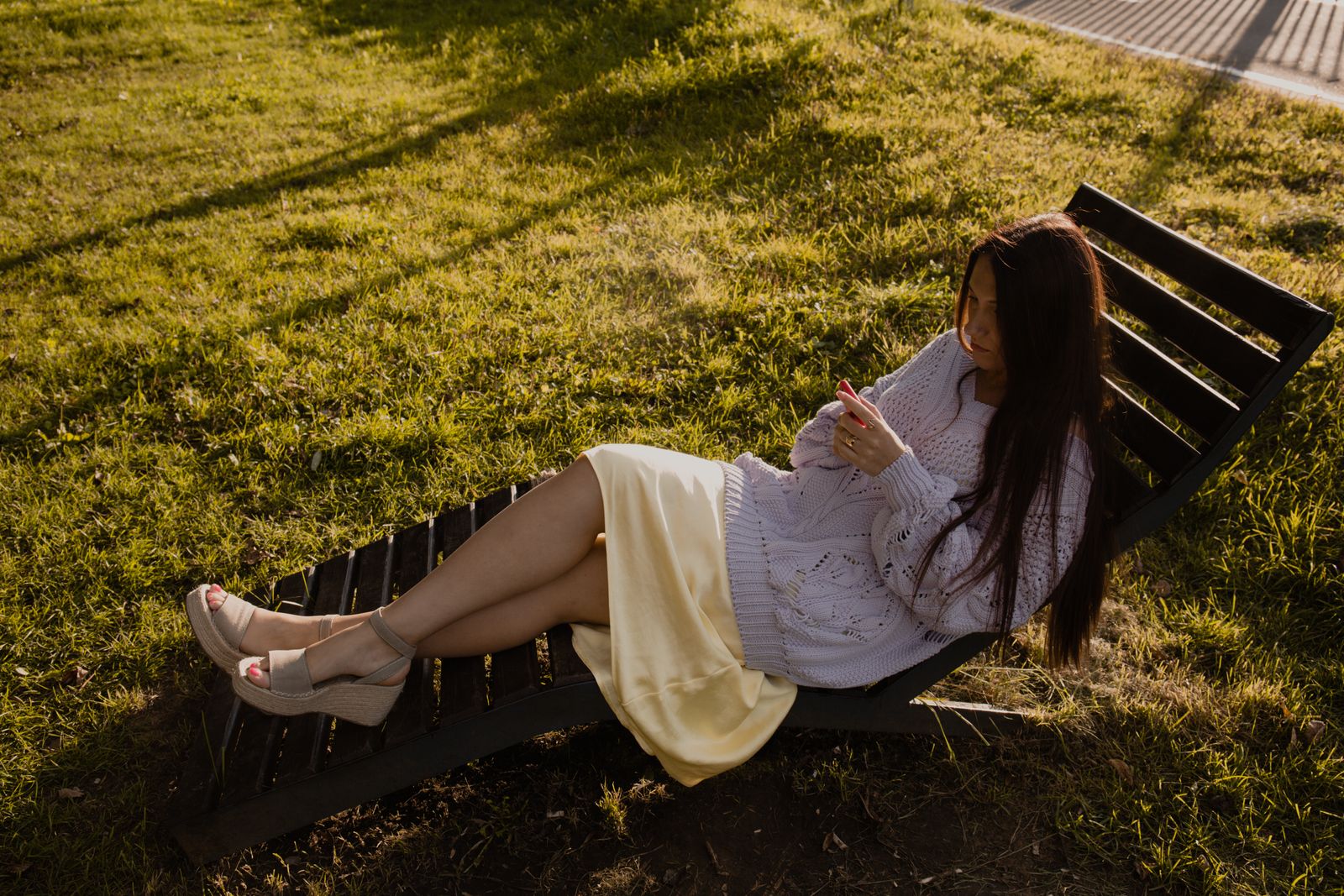 A woman in a sweater relaxes on a lounge chair in a grassy park, enjoying a sunny day.