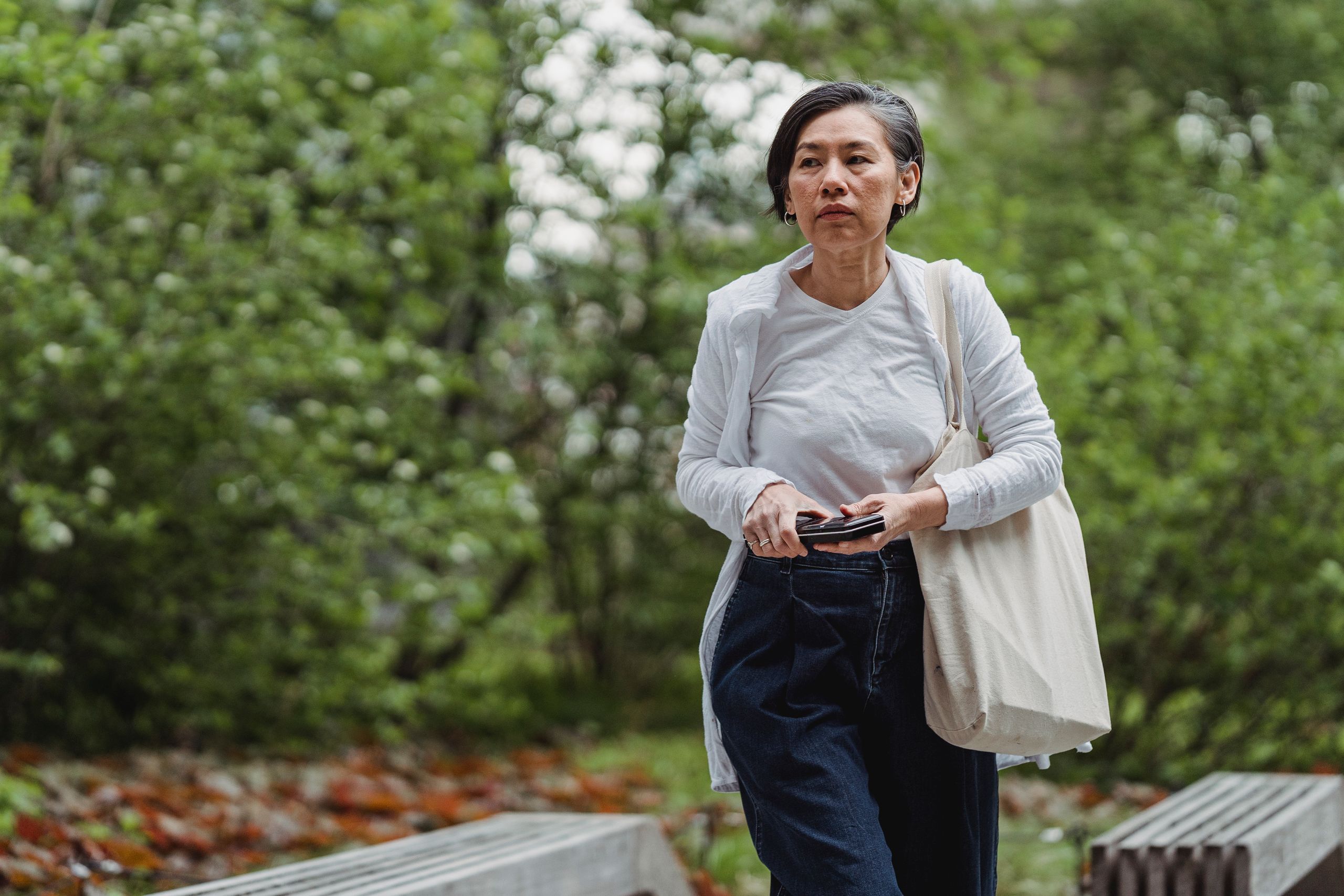 An adult woman walking outdoors with a phone and tote bag in hand. Casual and serene setting.