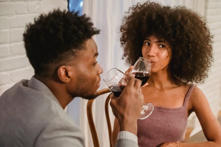 Thoughtful young African American couple in elegant clothes drinking red wine from glasses while sitting at table in chairs in bright house near curtains and brick wall