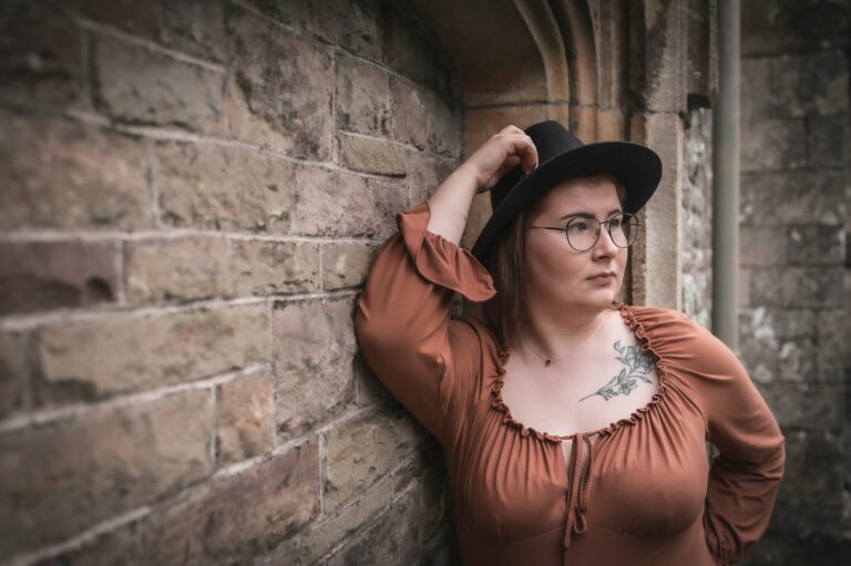 a woman wearing glasses and a hat leaning against a brick wall
