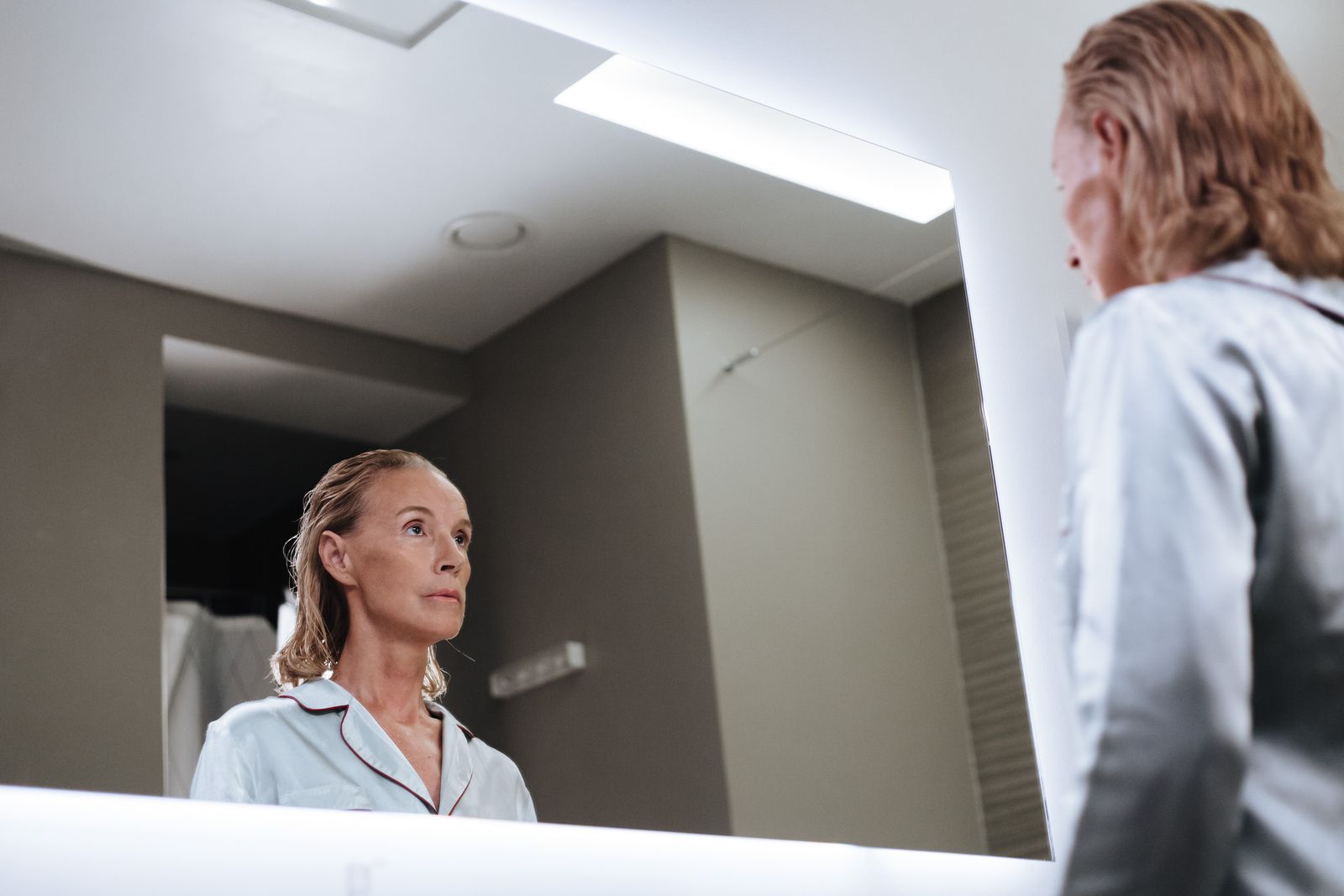 Senior woman looks introspectively at herself in a modern bathroom mirror.