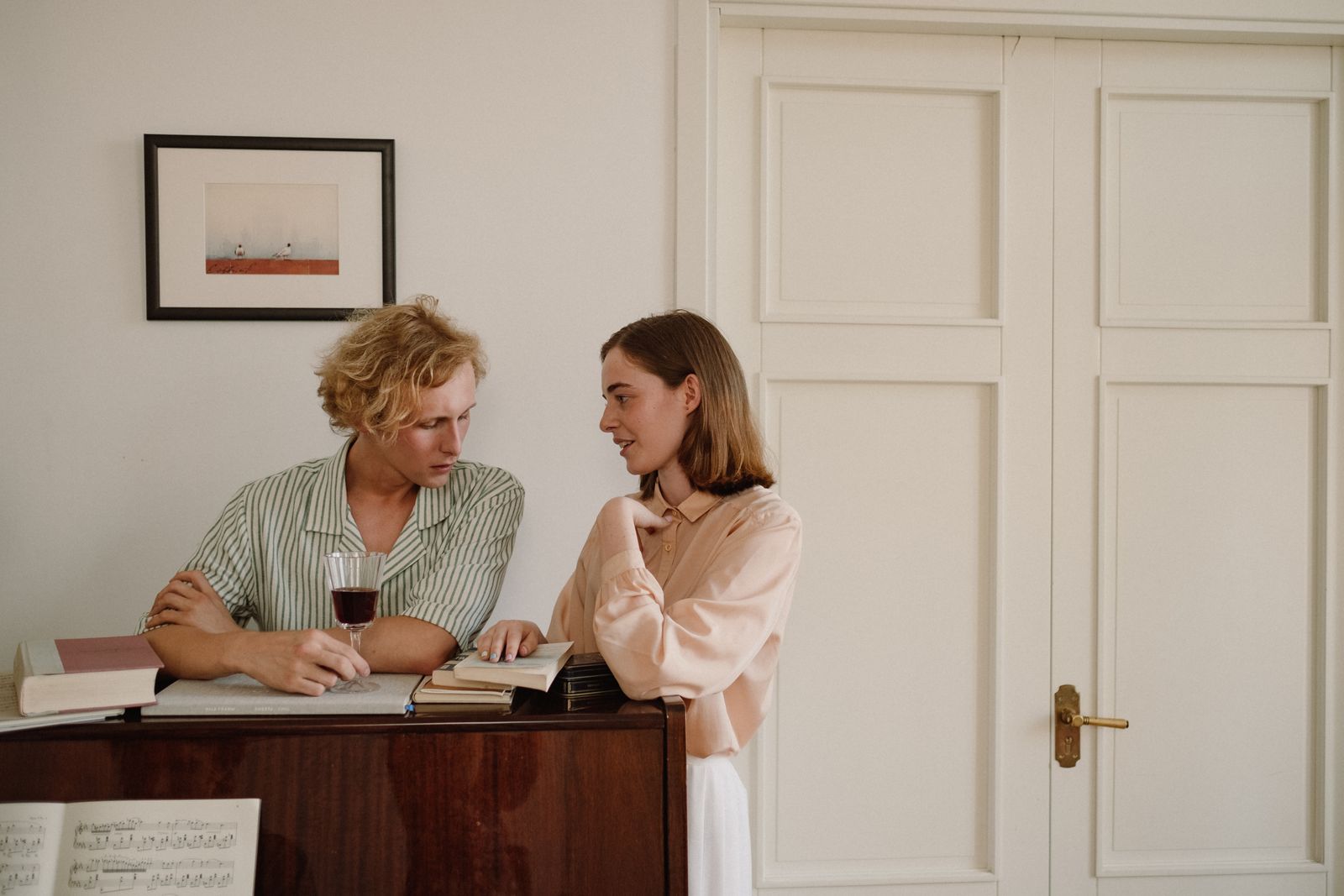 A couple engages in conversation by a piano, enjoying wine and books indoors.