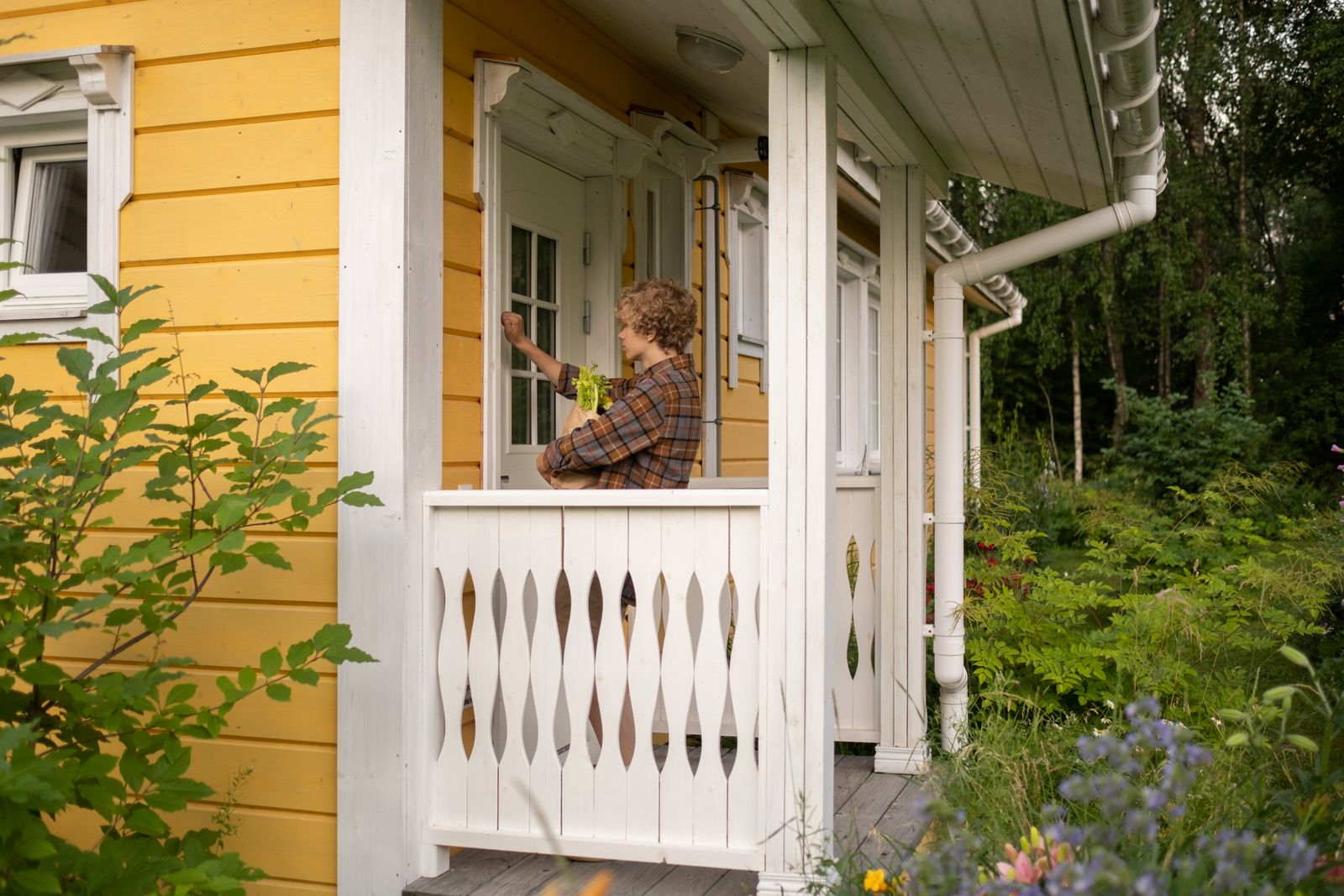 Curly-haired teenager knocking on the door of a yellow cottage surrounded by greenery.
