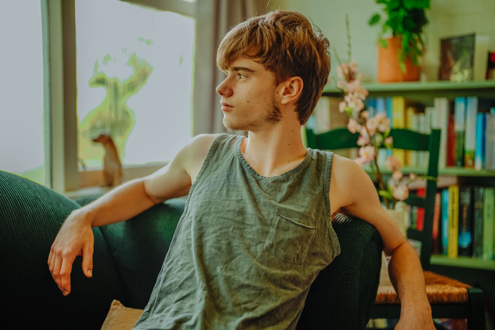 Young man in tank top sitting indoors, looking afar while relaxing by a bookshelf.