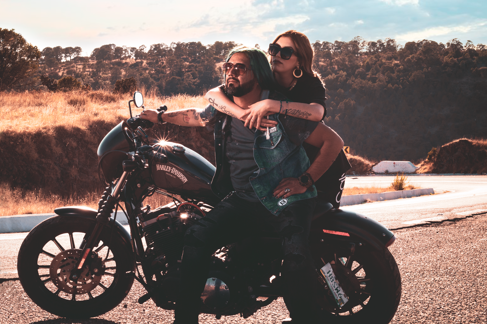 A couple embraces while sitting on a Harley Davidson motorbike on a scenic road.