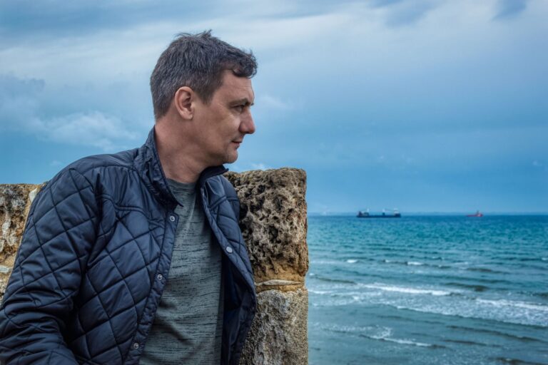 Middle-aged man in a jacket stands by ocean, deep in thought, on a cloudy day.