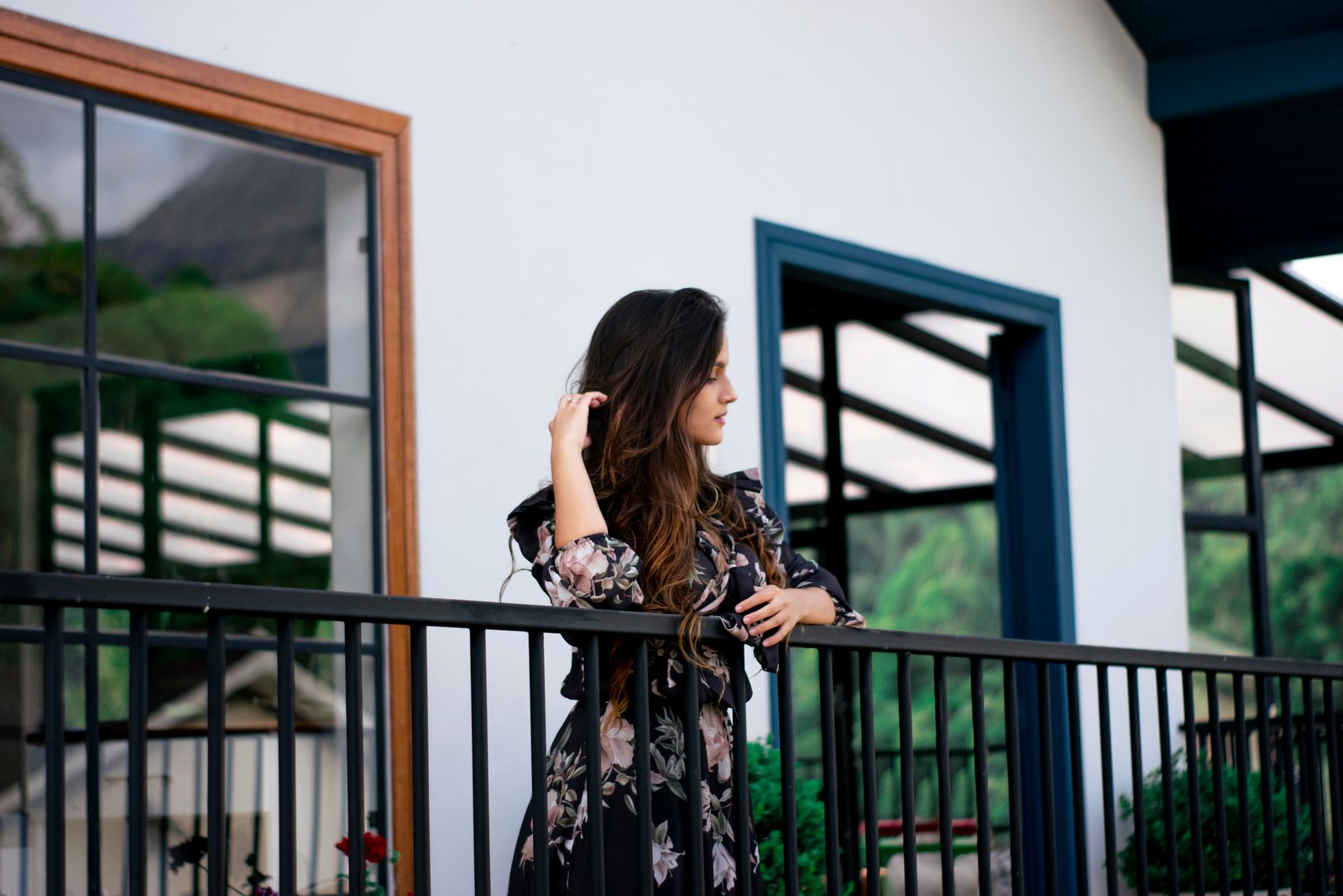 A woman in a floral dress leans casually on a balcony railing of a modern house.
