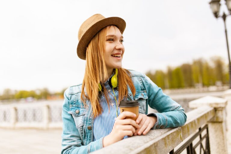 Smiling young woman wearing a hat and denim jacket, enjoying a coffee outdoors during a sunny spring day.