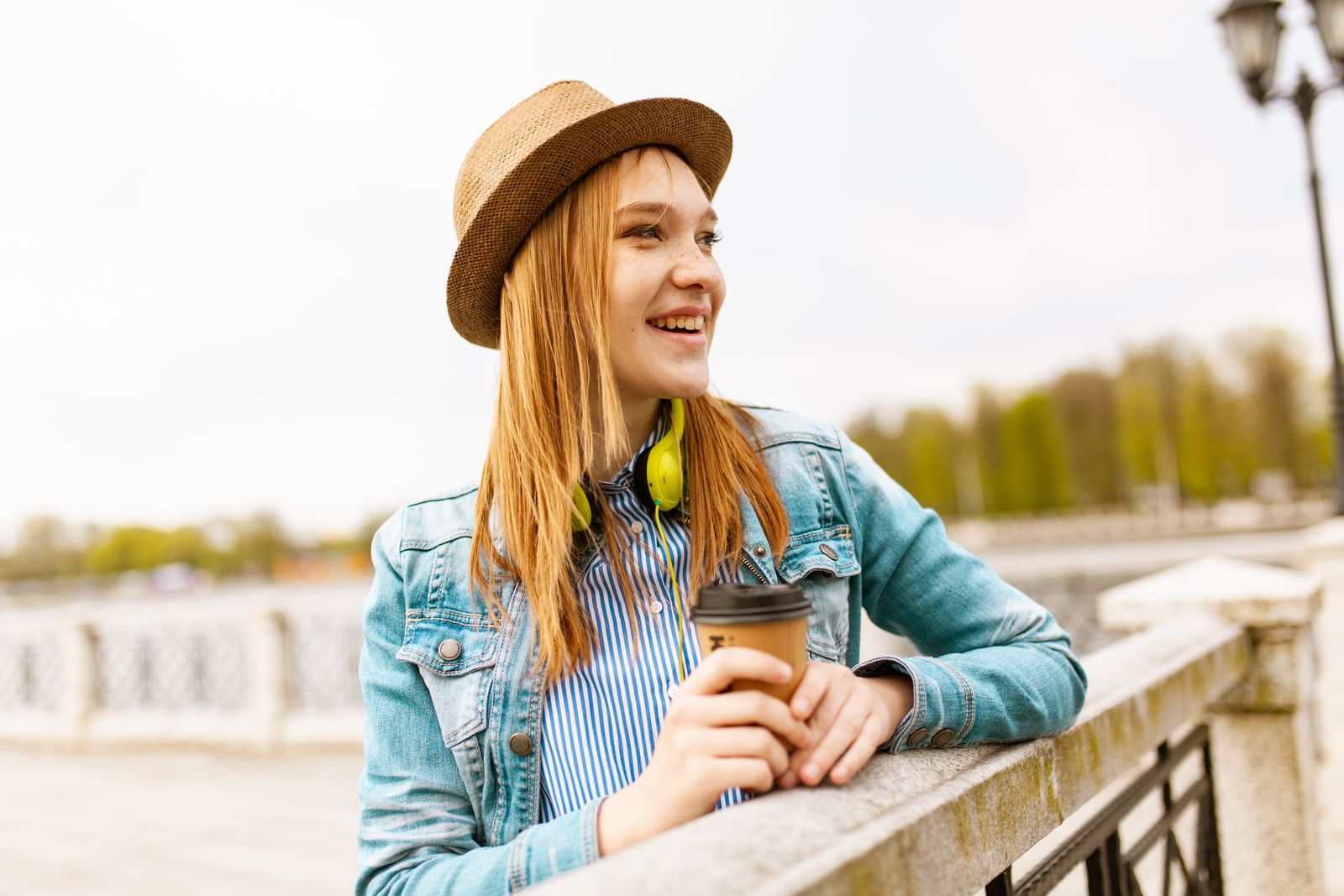 Smiling young woman wearing a hat and denim jacket, enjoying a coffee outdoors during a sunny spring day.