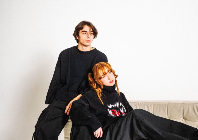 A fashionable couple dressed in black sits and poses thoughtfully on a couch.