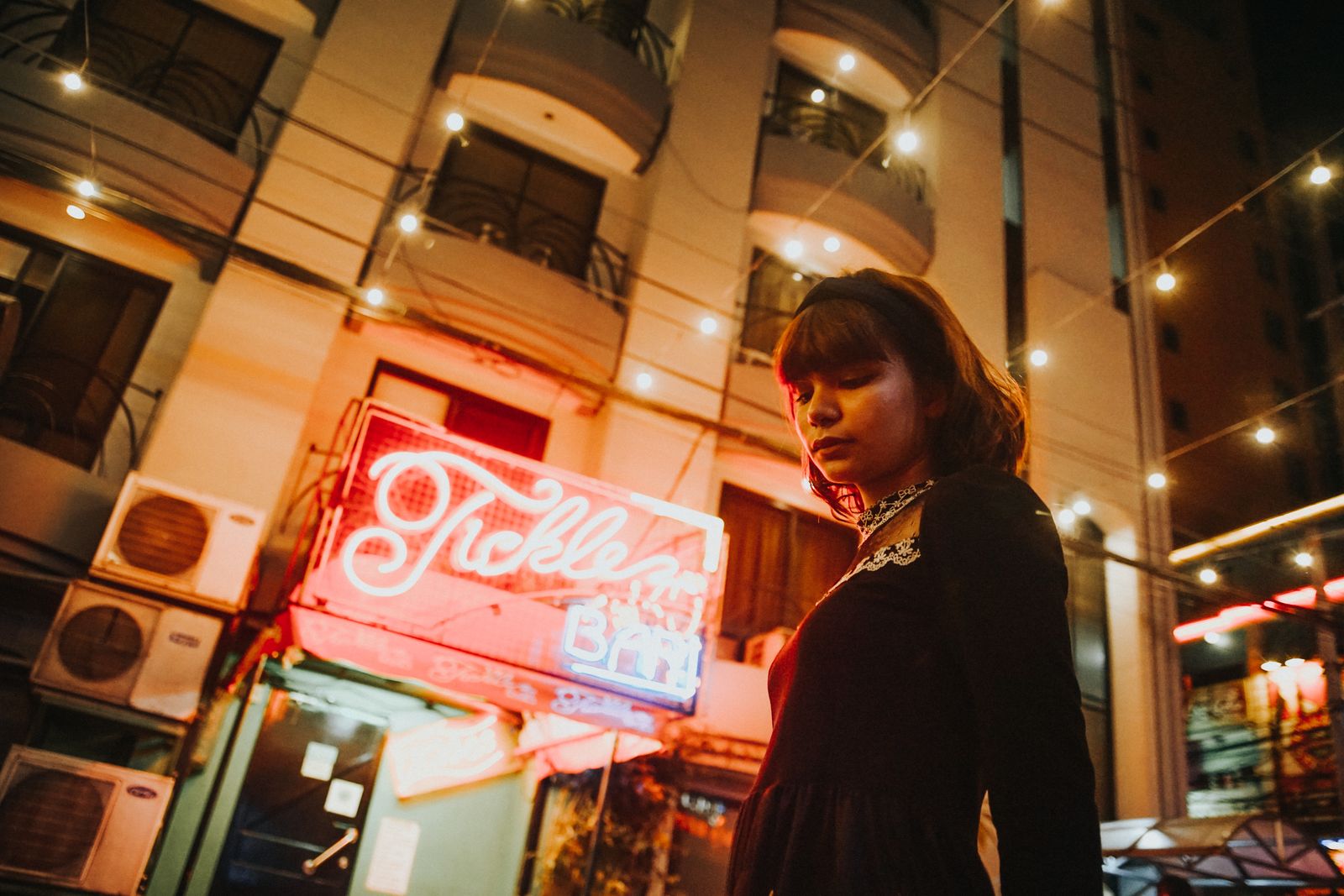 A woman stands under a neon sign in a city, evoking urban nightlife vibes.