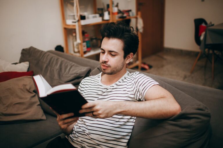 Man in a striped shirt relaxing on a sofa, reading a book indoors.