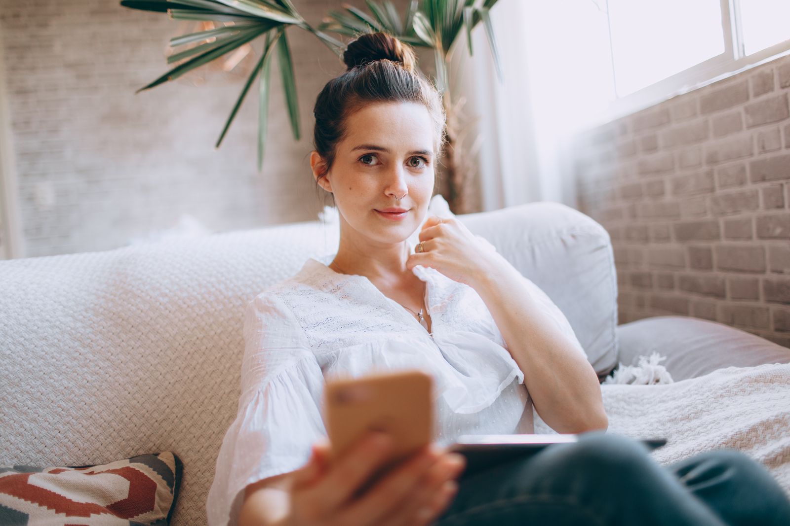 Young woman holding smartphone indoors, relaxing on sofa in bright living room.
