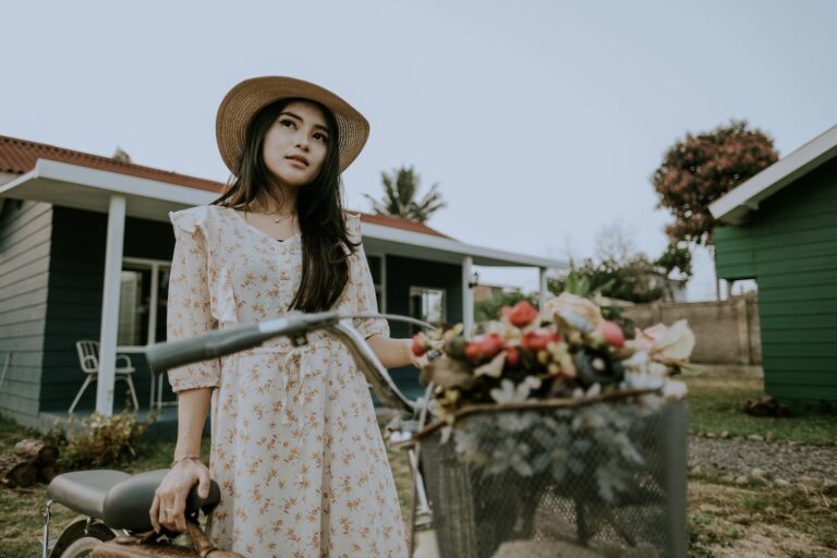 A stylish woman in a summer dress stands with a bicycle in a garden setting.