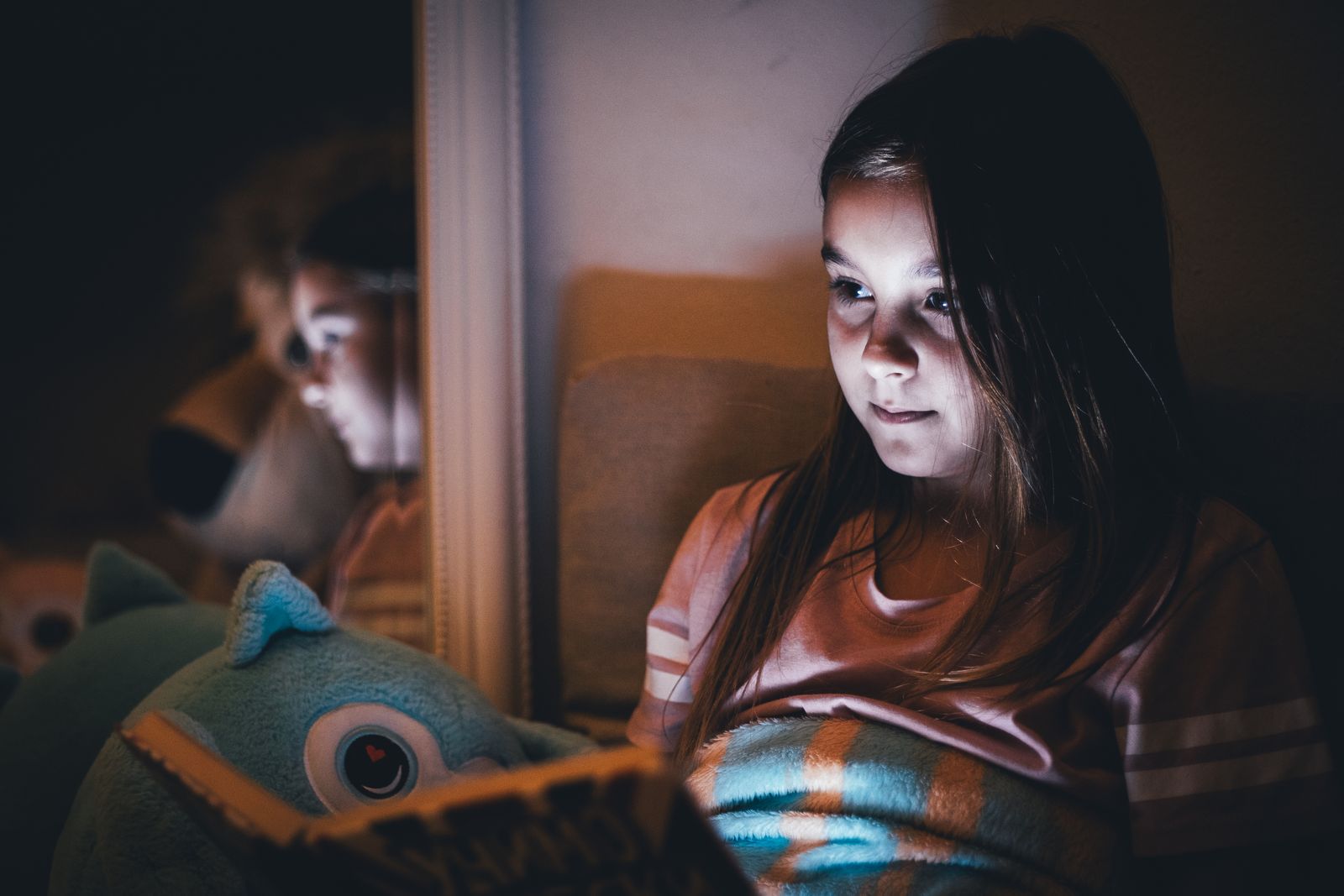 A young girl reads a book under warm light at night, reflecting on a mirror.