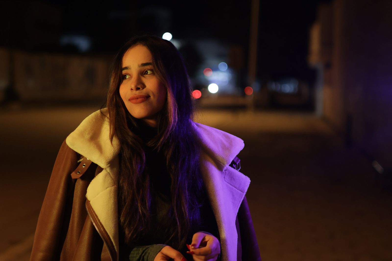 A young woman in a warm coat smiles on a city street at night, capturing a vibrant urban vibe.
