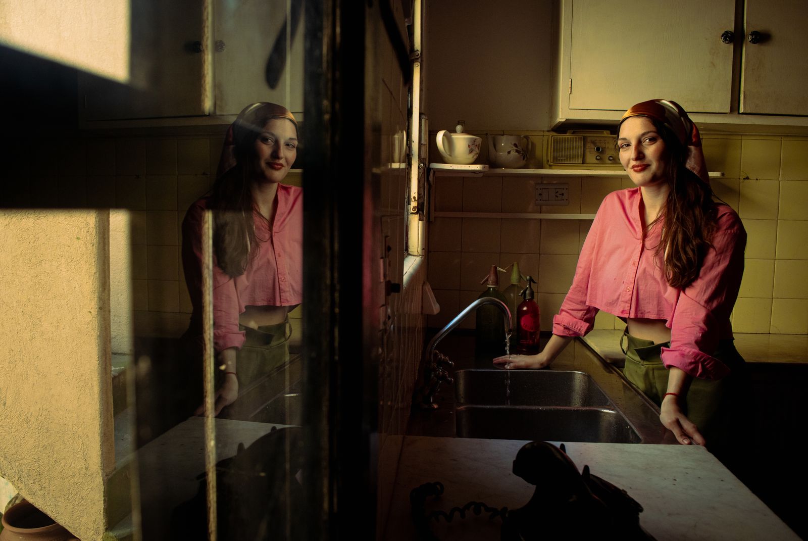 A woman in a vintage pink blouse and headscarf stands by a kitchen sink with sunlight cast on her face.