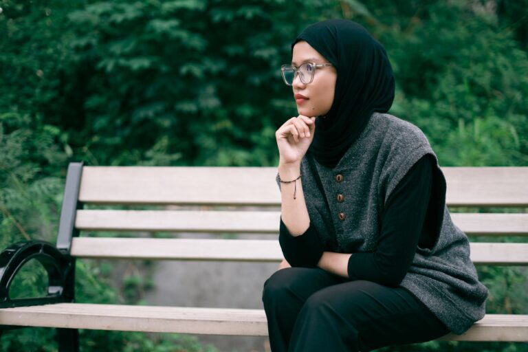 Young woman in hijab sitting on a wooden bench in a lush green park, deep in thought.