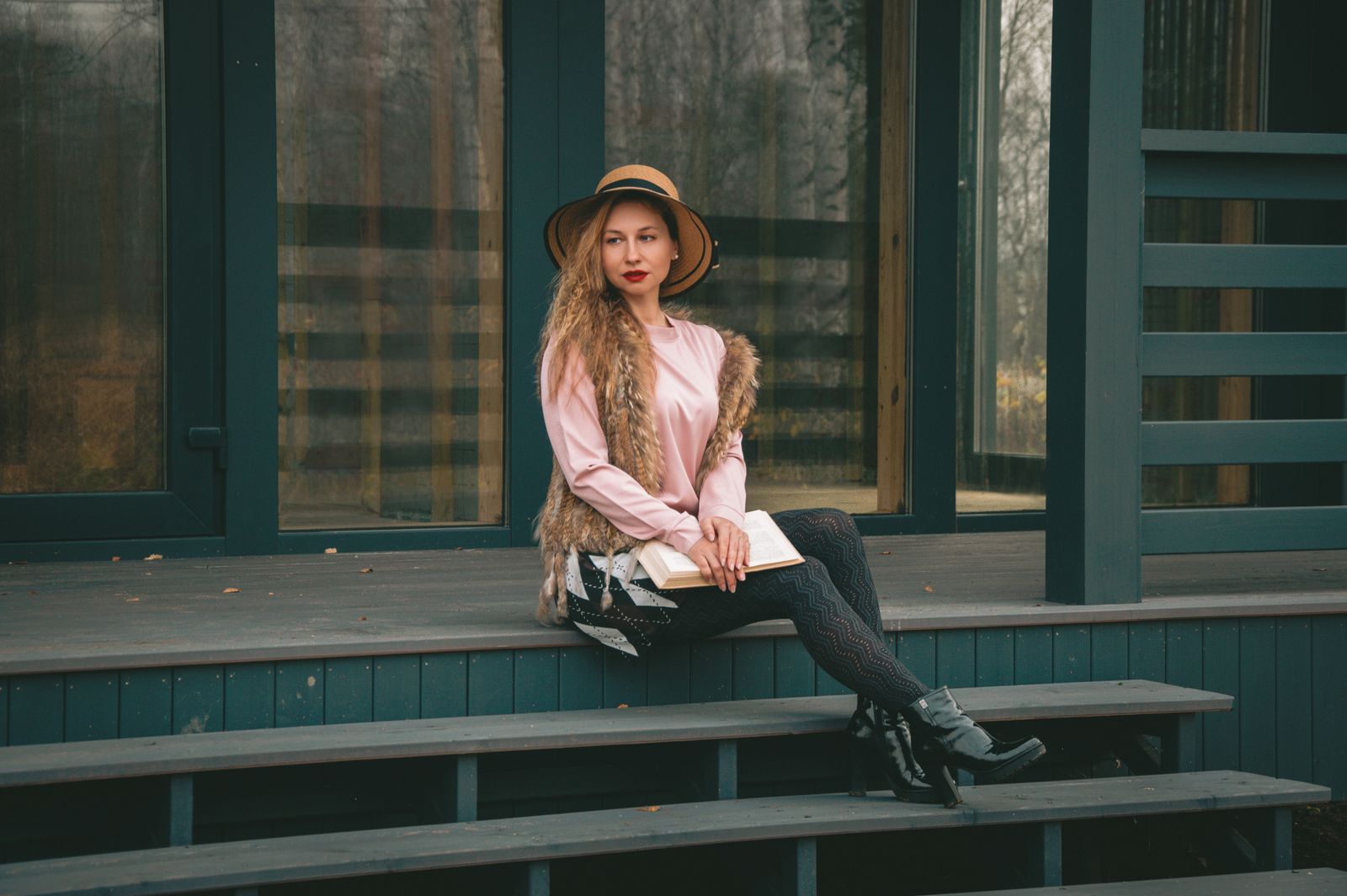 Woman in fashionable attire seated on porch steps, holding a book, wearing a hat and fur vest.