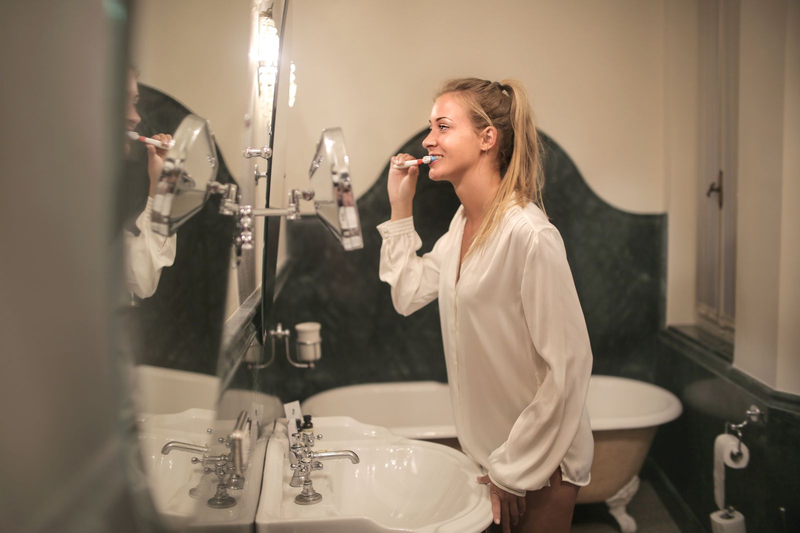 Side view of blonde female with ponytail in blouse standing in bathroom while brushing teeth and looking at mirror