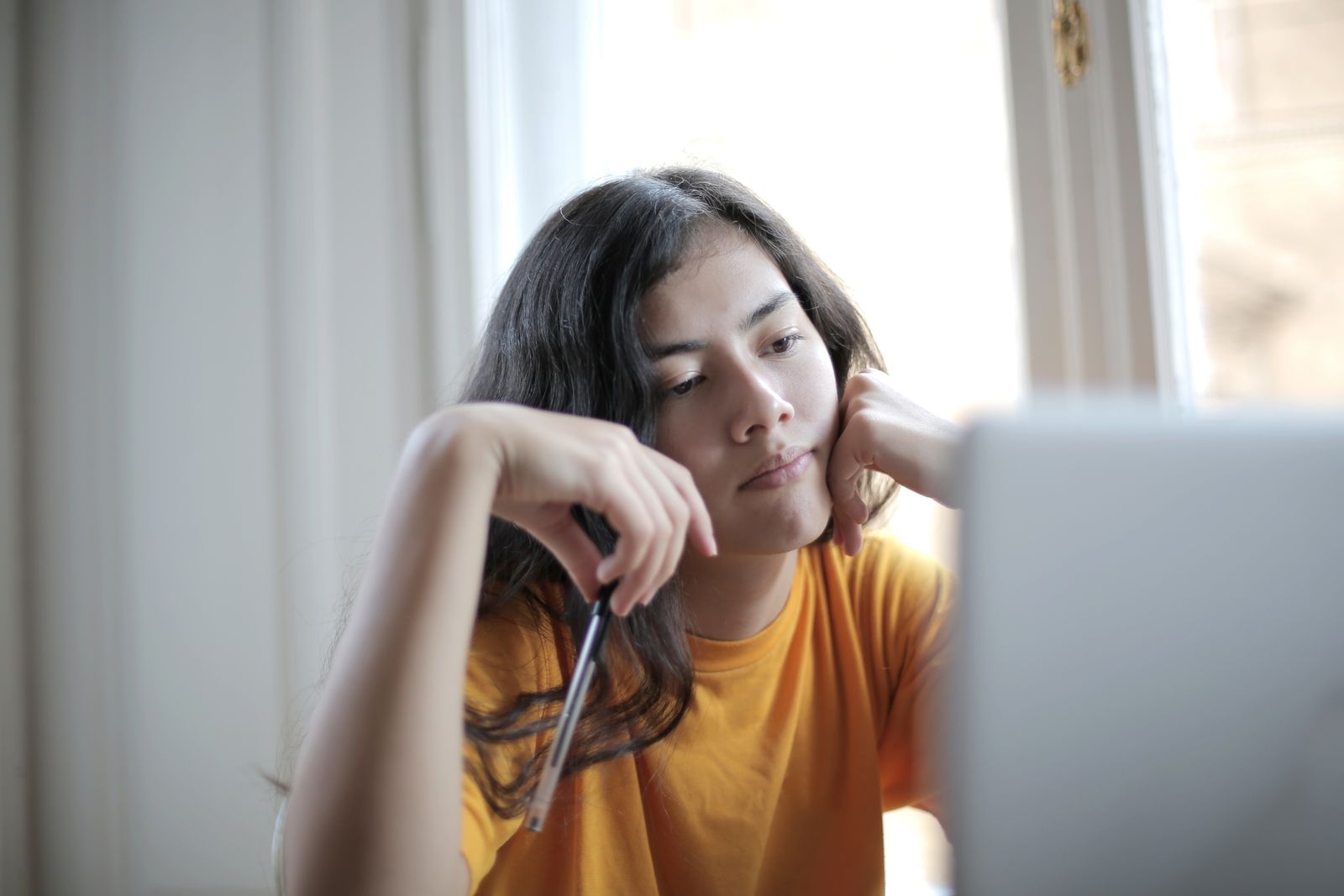 Pensive woman in a yellow shirt thoughtfully holding a pen near a window indoors.