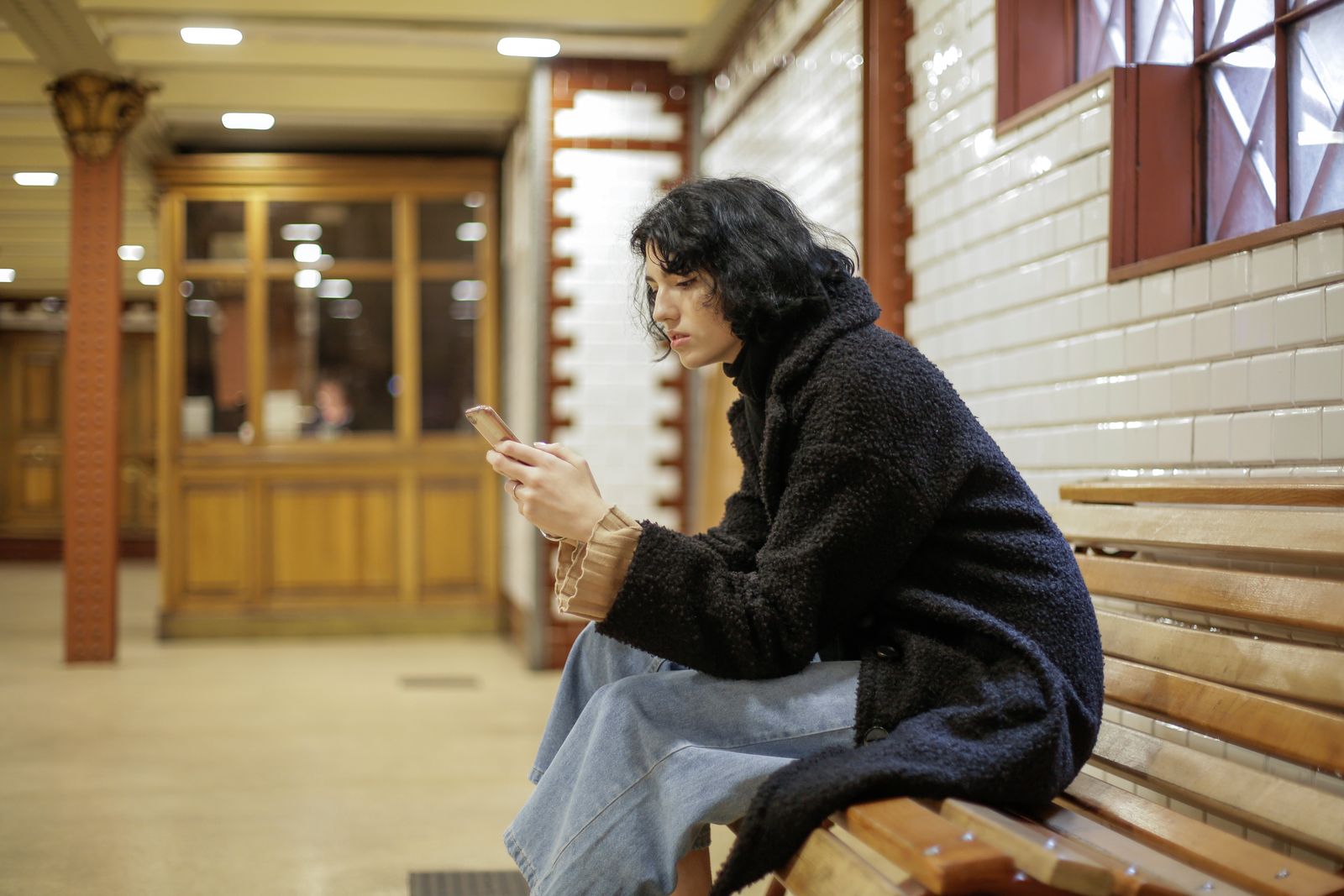 A young woman in a black coat sits on a bench using her smartphone in a well-lit indoor setting.