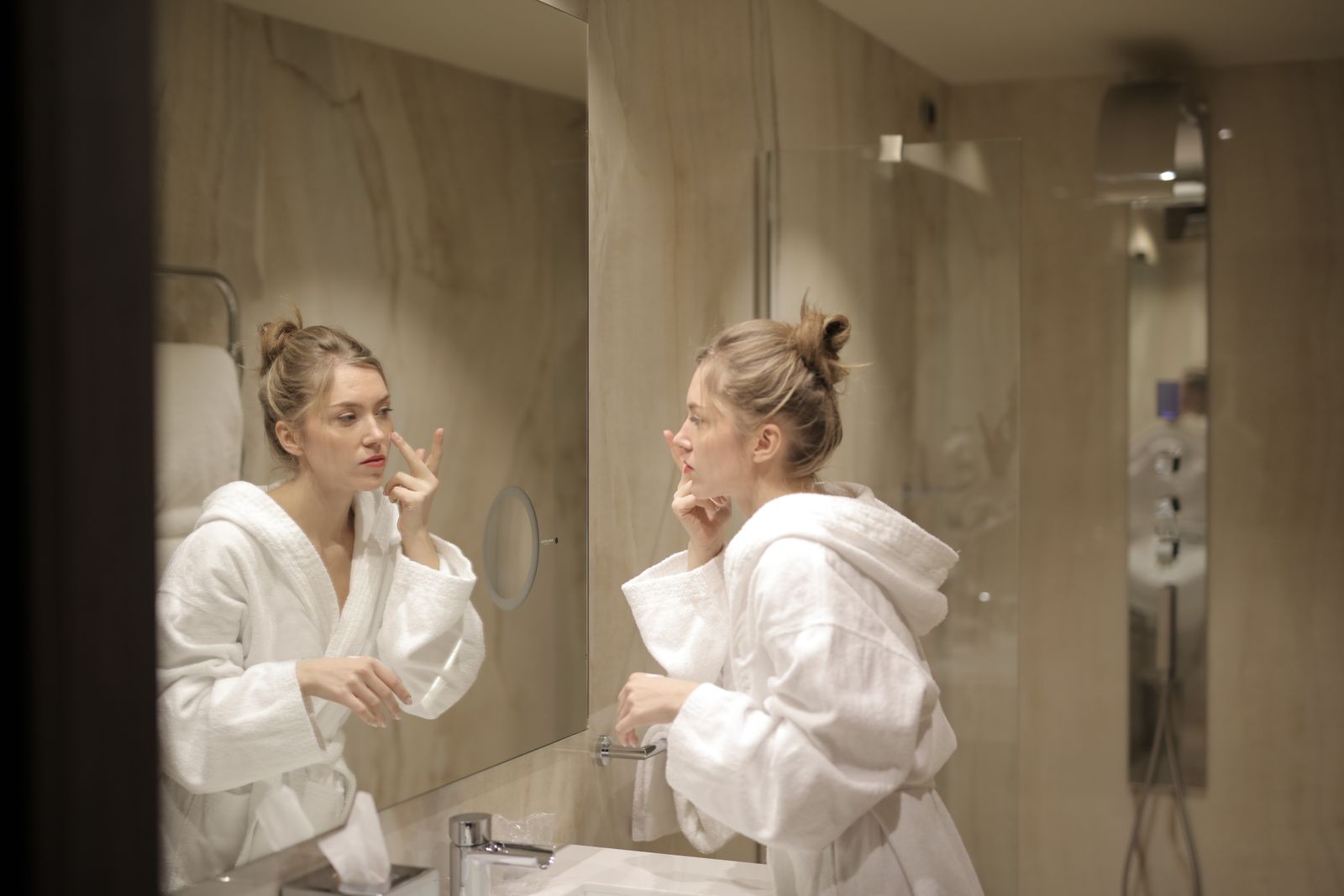 A woman in a white bathrobe applies moisturizer while looking at her reflection in the bathroom mirror.