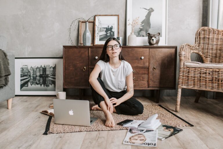 Dream young woman in trendy casual wear sitting with crossed legs on floor near chest of drawers and thinking while resting at home