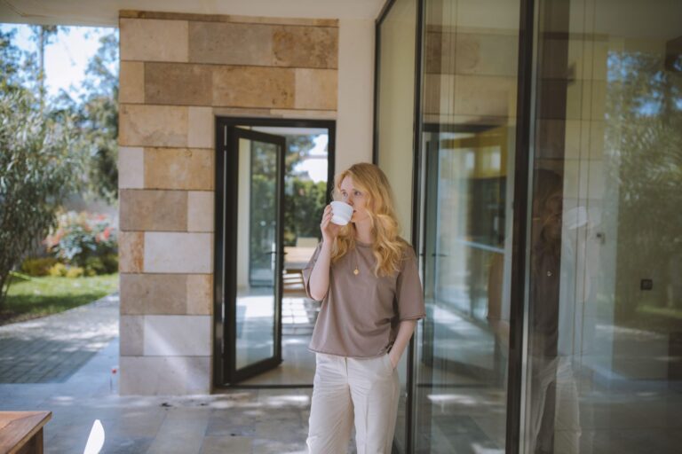 Blonde woman sipping coffee near glass wall outdoors. Relaxed atmosphere.