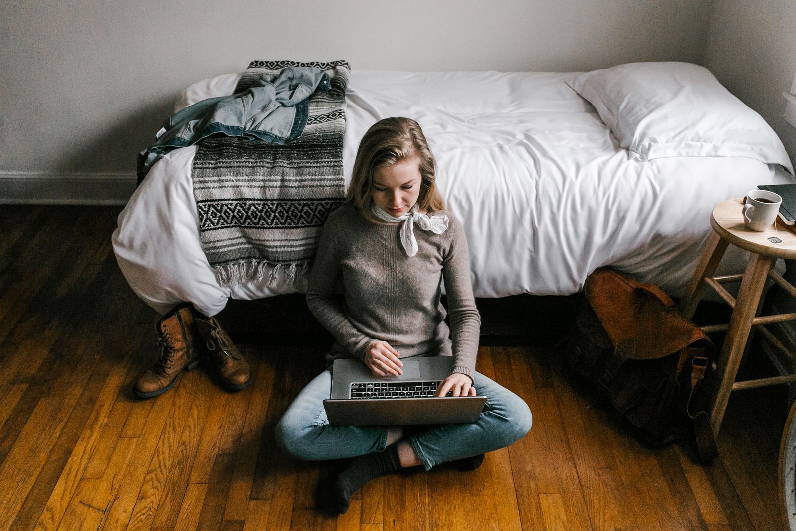 A young woman comfortably working on a laptop in a modern, minimalist bedroom setting.