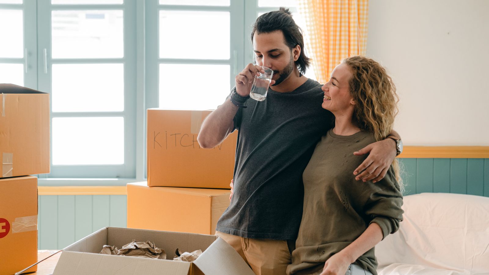Ethnic man drinking water while cuddling happy girlfriend standing near pile of cardboard boxes in sunlight after moving into new apartment