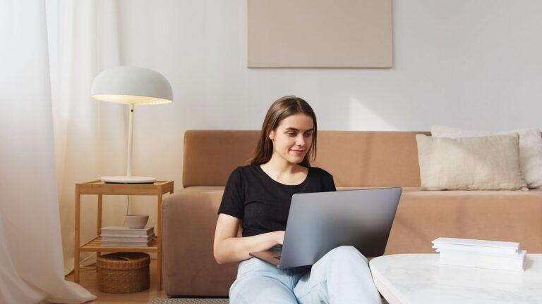 Happy content young female in casual clothes sitting on floor near comfy sofa and browsing modern netbook in cozy living room