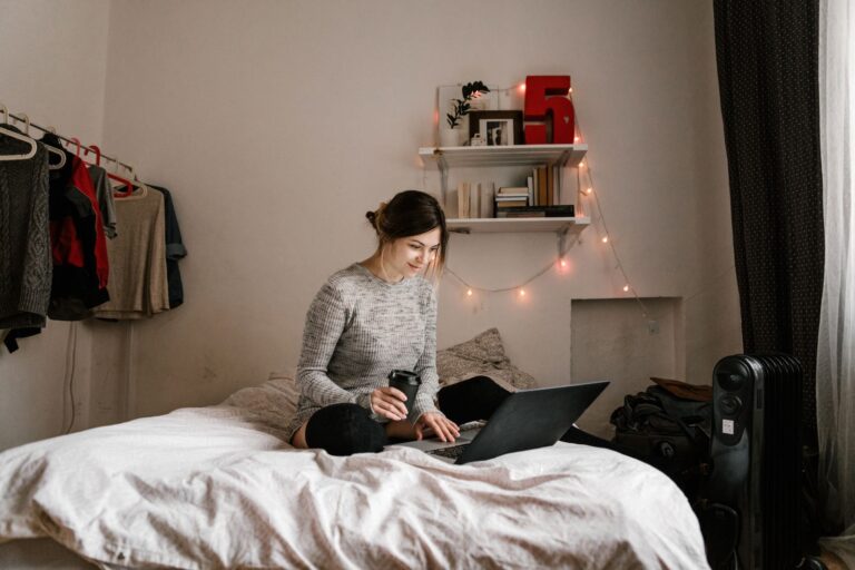 Young woman enjoying coffee while using a laptop in a cozy, well-lit bedroom setting.