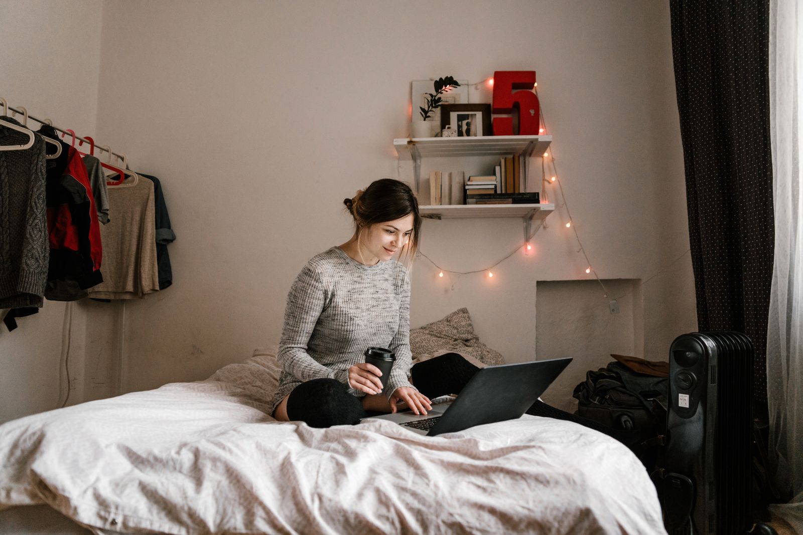 Young woman enjoying coffee while using a laptop in a cozy, well-lit bedroom setting.